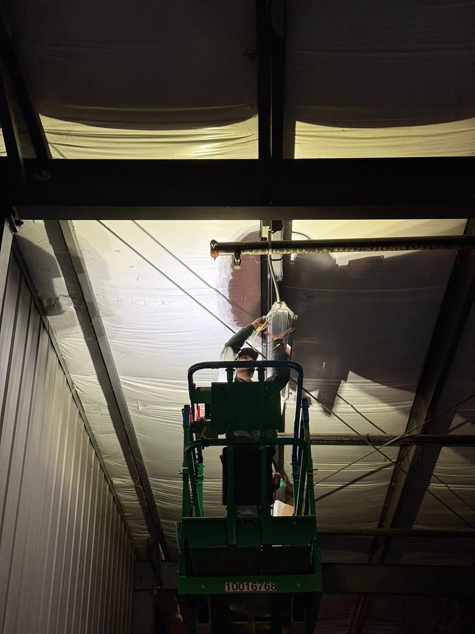 A person in a green scissor lift works on the ceiling of a large industrial warehouse.