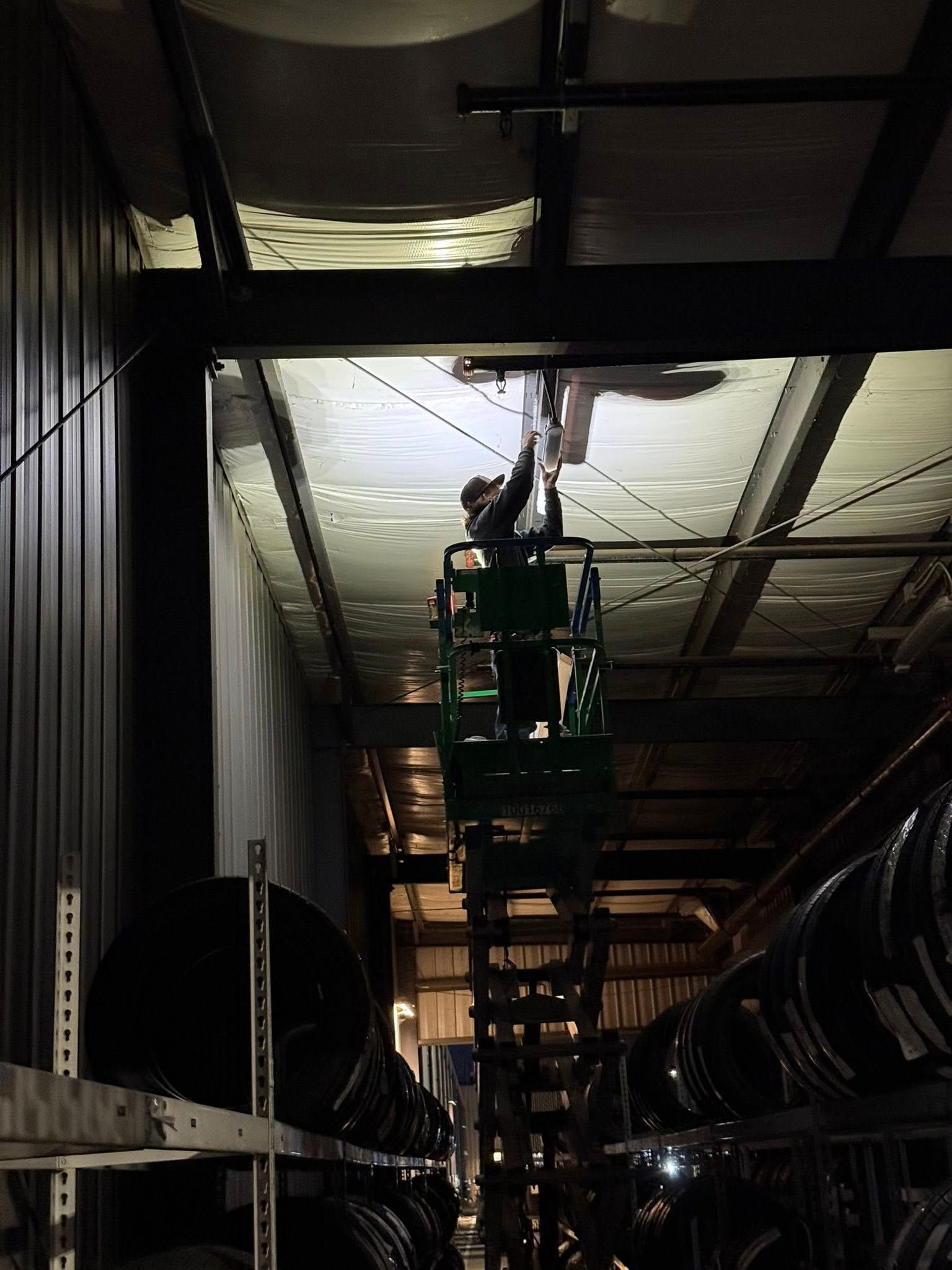 A worker on an elevated scissor lift works on the metal ceiling of a dark warehouse lined with stacked industrial barrels.