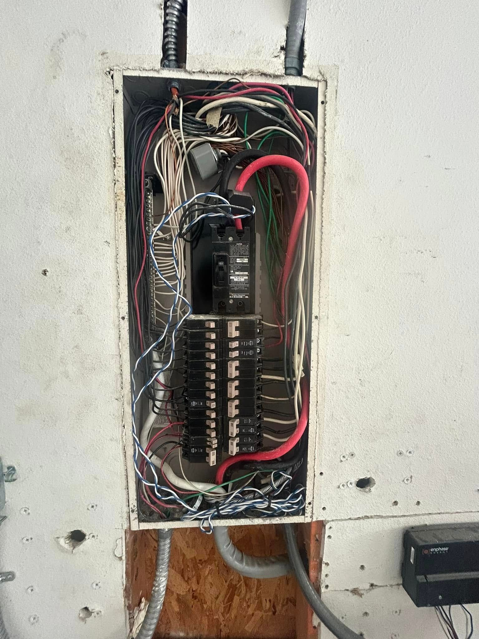 An open electrical breaker panel showing wires, circuit breakers, and conduit on a wall.