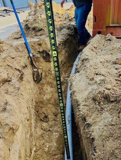 A measuring rod placed in a narrow trench next to a pipe, with a person standing nearby.