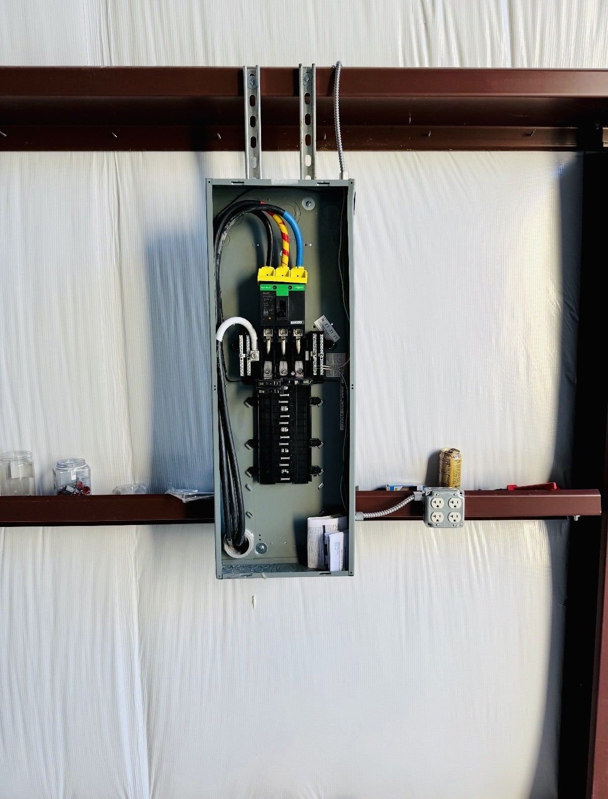 An open electrical breaker panel mounted on a metal wall, with wires connected to breakers and a side junction box.