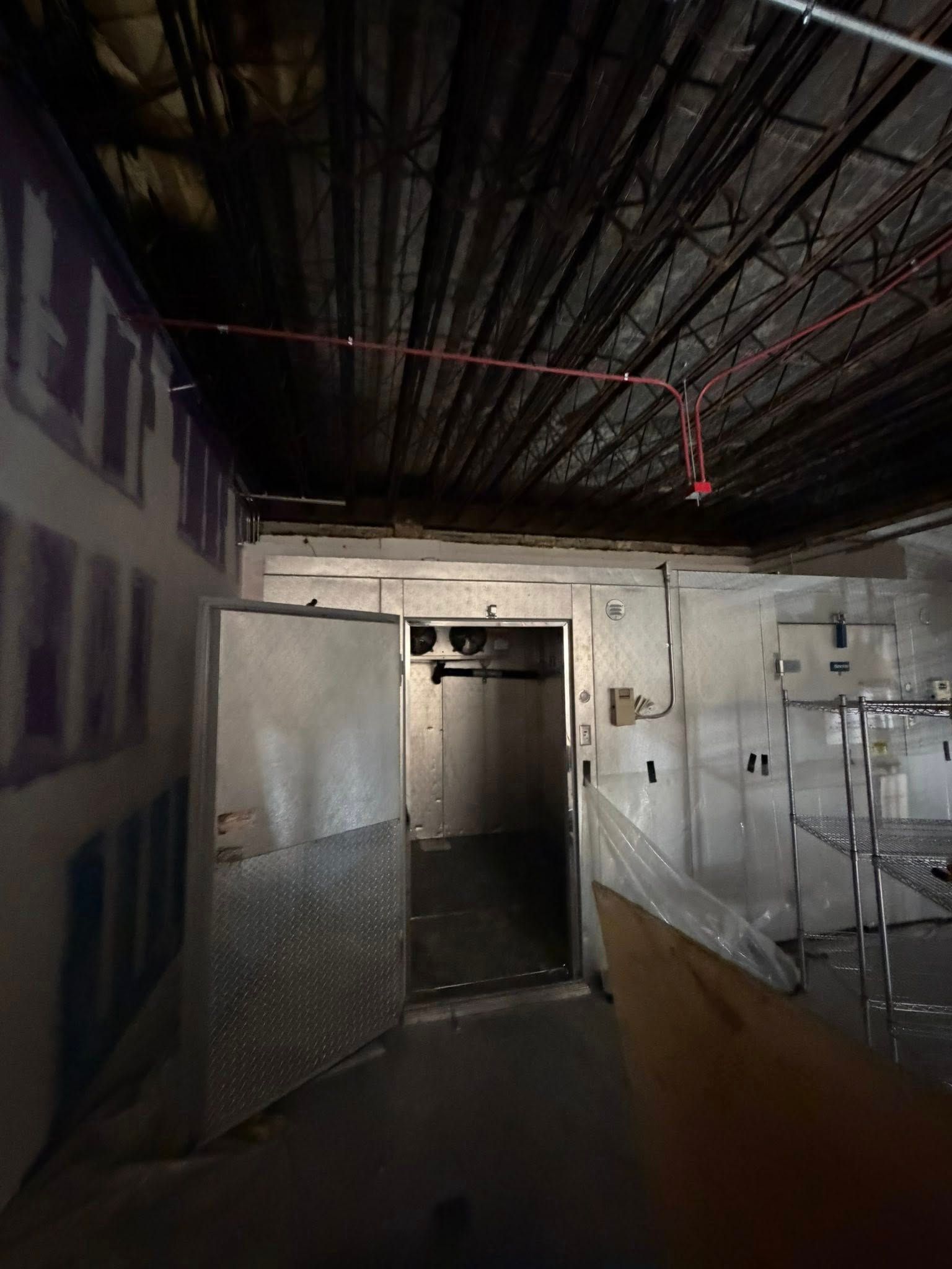 A metal walk-in cooler door stands open in an unfinished room with an exposed ceiling and industrial metal shelving.