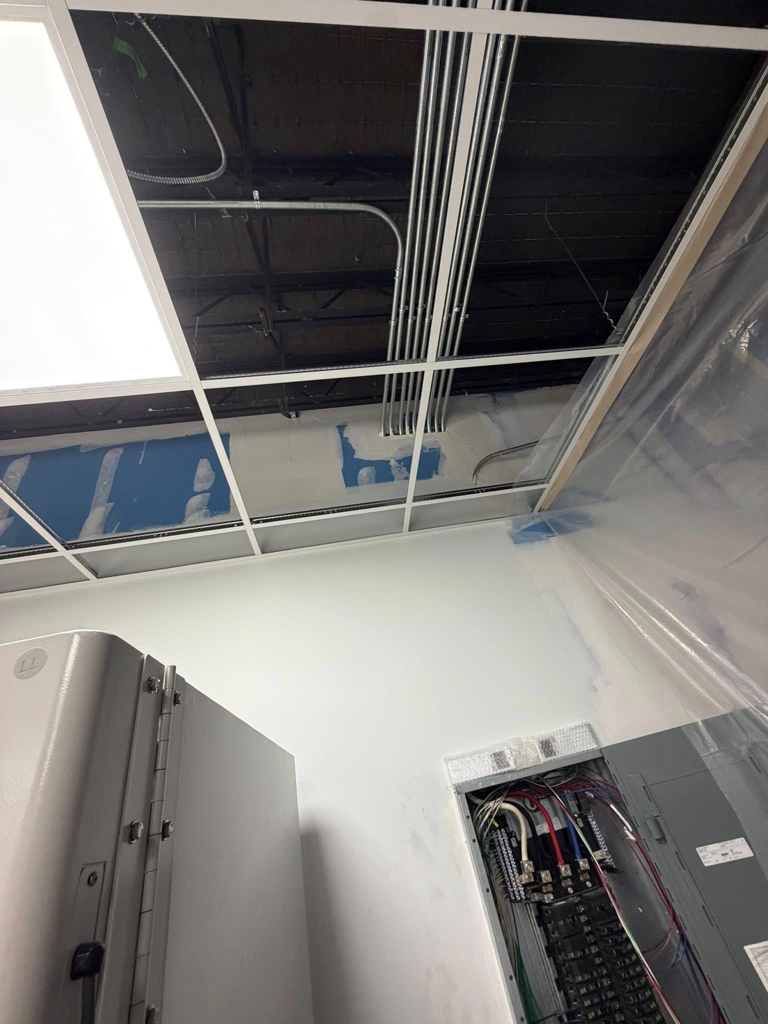 A low-angle view of a drop ceiling under construction with exposed wiring, metal conduit, and an open electrical panel.