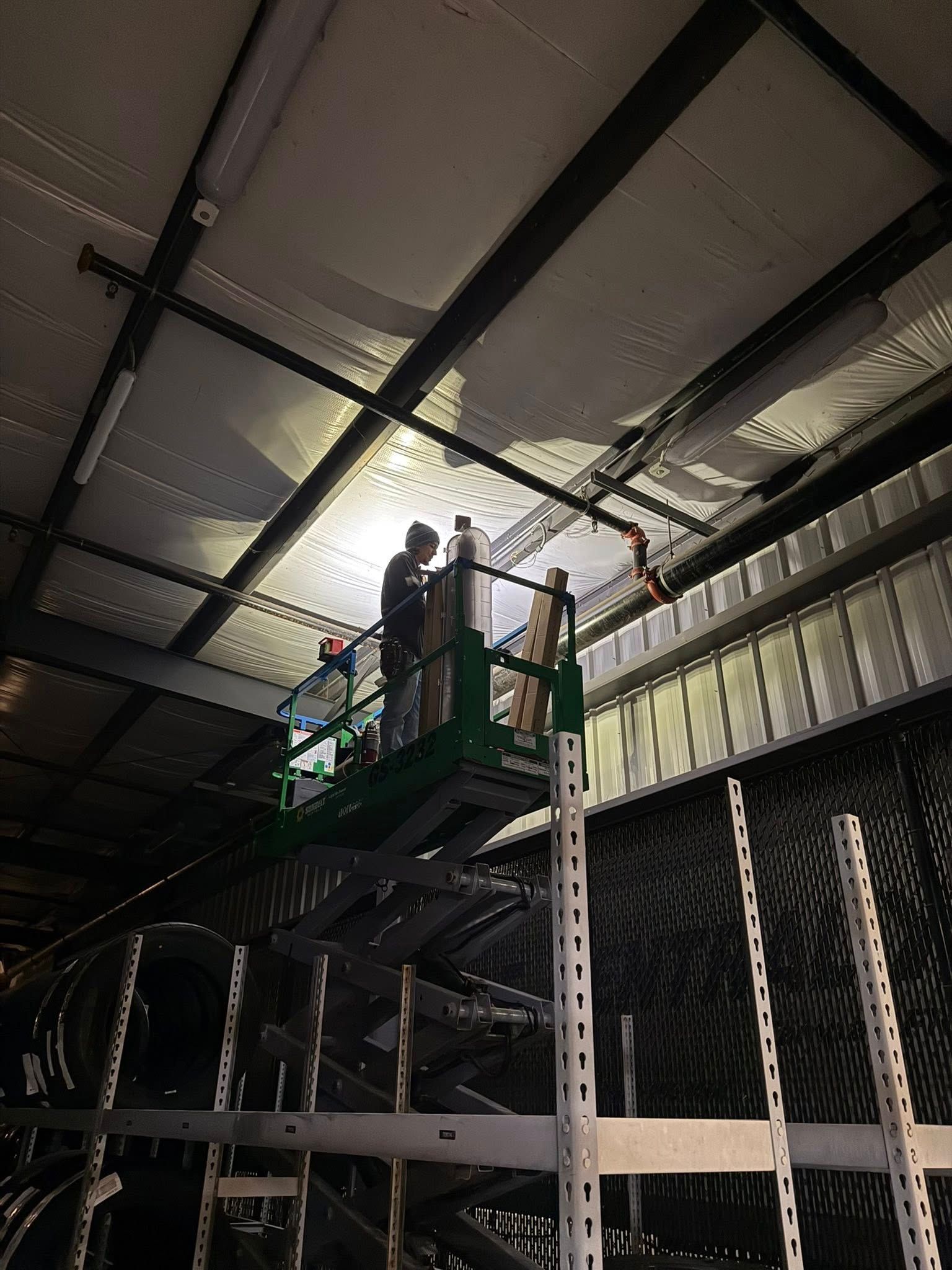 A person working on a green scissor lift in a warehouse, reaching toward the ceiling near metal racking.