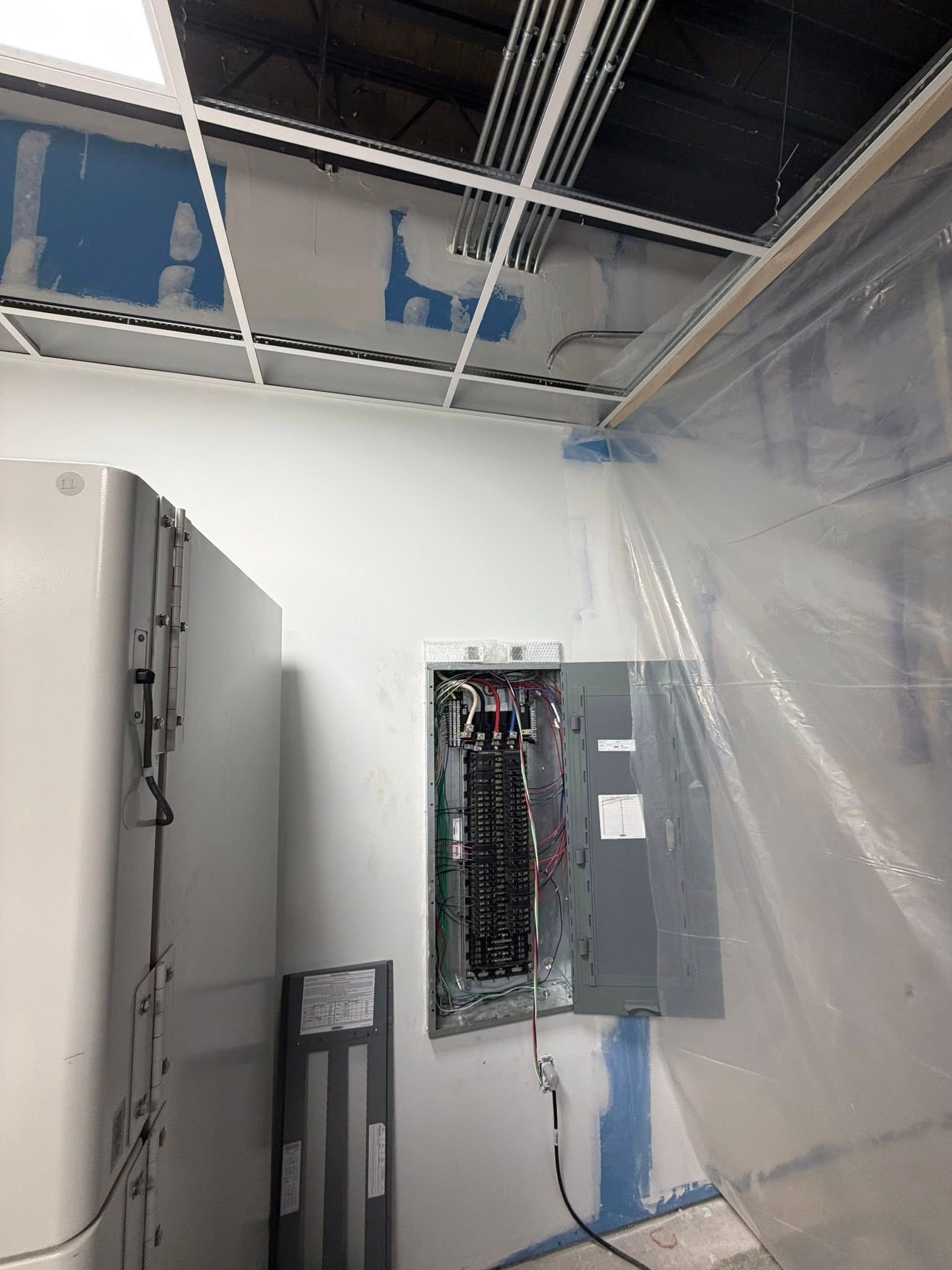 An open electrical panel mounted on a wall in a room with exposed ceiling grids and plastic sheeting.