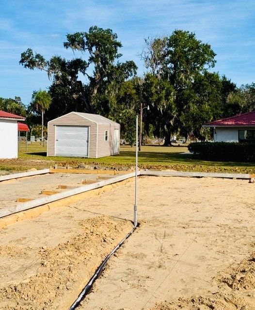 Construction site showing a leveled sandy lot with concrete footings, a vertical pipe, and a shed in the background.