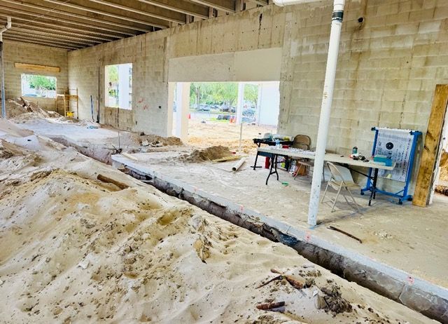 Interior of a building under construction, showing a sandy floor with a trench, concrete walls, and a temporary workbench.