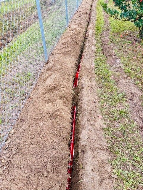 A narrow trench runs along a fence line, containing a red irrigation pipe laid in the soil.