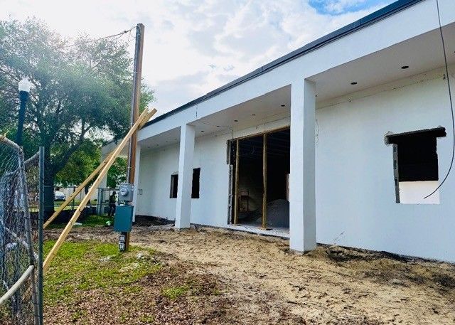 A white building under construction with a large open doorway, a covered porch, and a dirt yard with a utility pole.