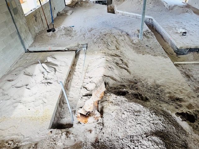 An unfinished interior construction site with sand floors, concrete foundation steps, and exposed plumbing pipes.