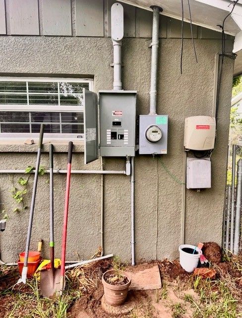 An electrical service panel and meter mounted on an exterior stucco wall, with gardening tools leaned against it.