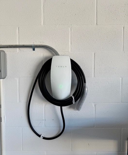 A white Tesla wall connector mounted on a white cinder block wall with a black charging cable coiled around it.