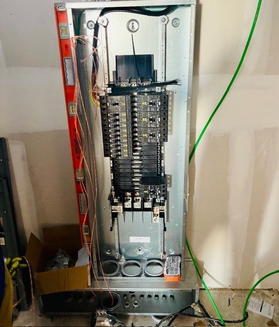 An open electrical service panel showing breakers, wiring, and conduit entries on a wall in a room under construction.
