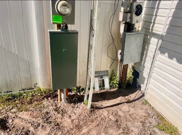 An exterior electrical service setup with a meter, a large green enclosure, and a gray box mounted on a wall near sand.