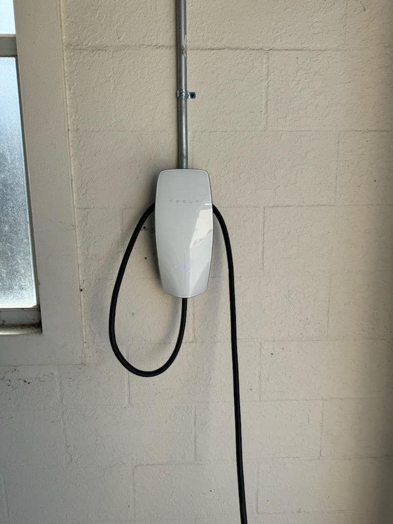 A white Tesla wall connector mounted on a light-colored brick wall with a black charging cable hanging from it.