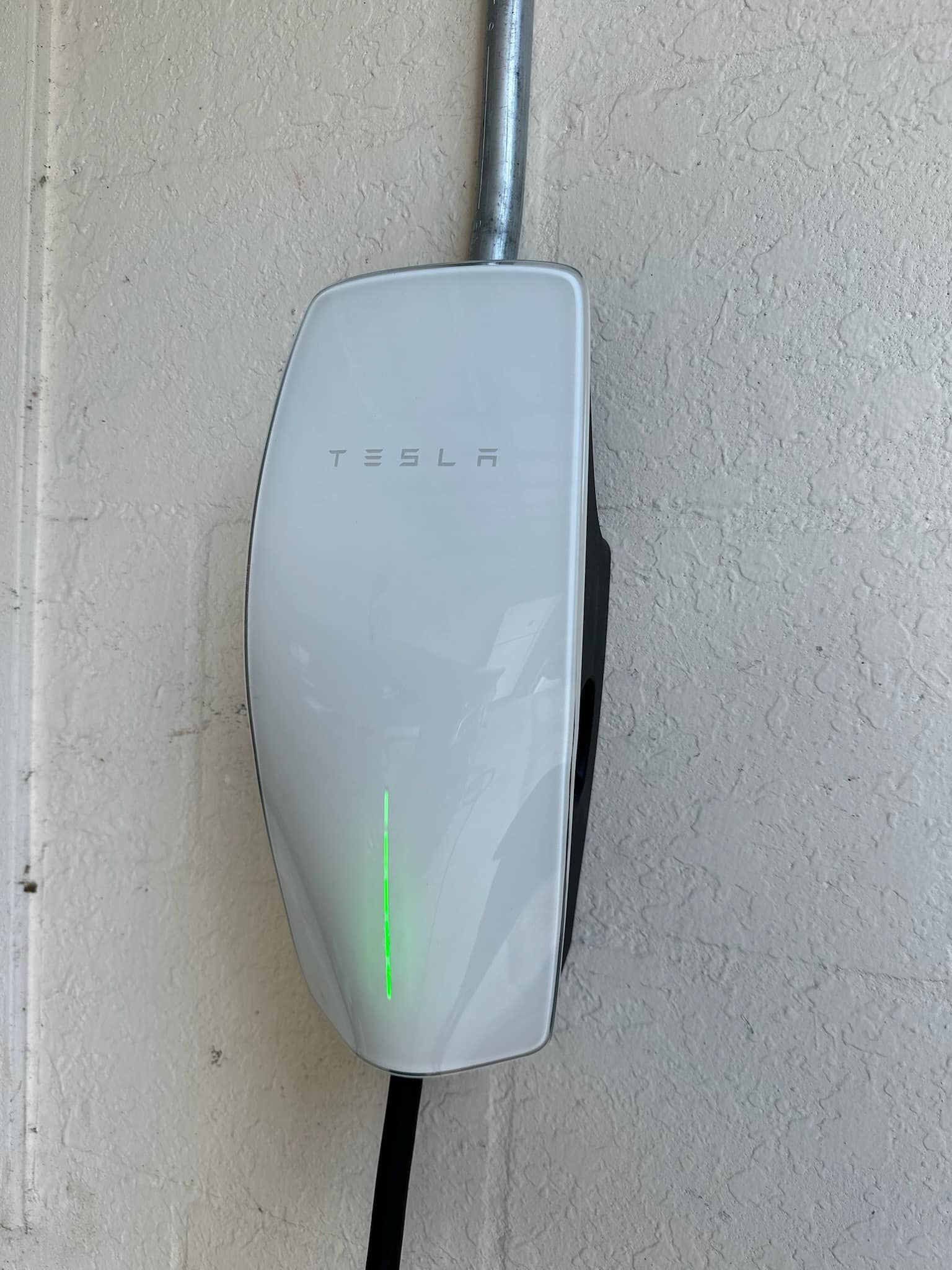 A white Tesla wall charger mounted on a textured wall with a glowing green status light.