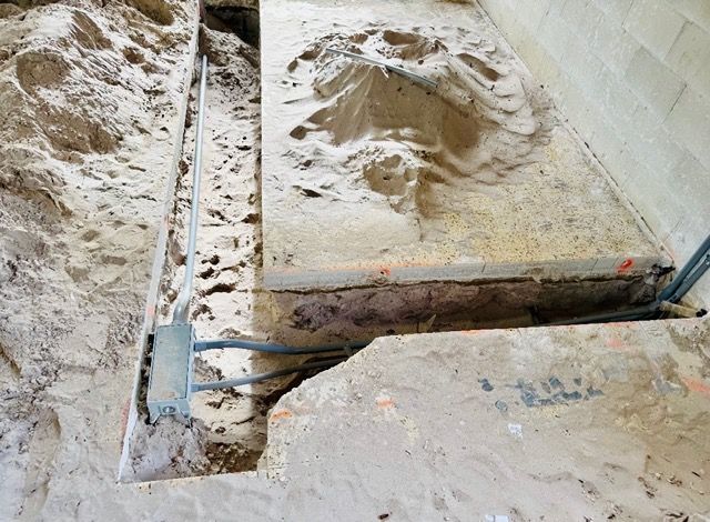 A construction trench in sand with a metal electrical junction box and conduit pipes running along the concrete slab edge.