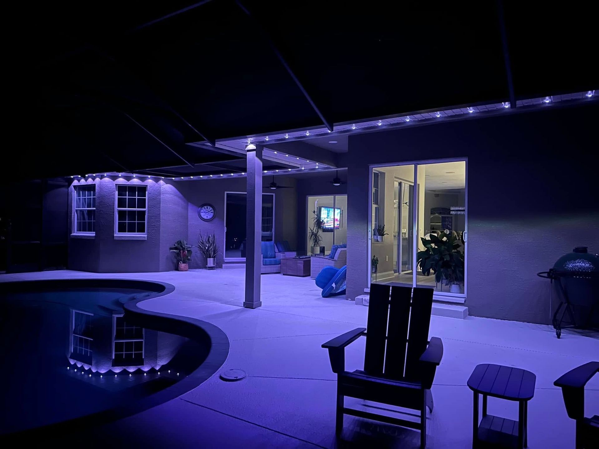 A patio at night with purple LED lights along the roofline, reflecting in a swimming pool next to Adirondack chairs.
