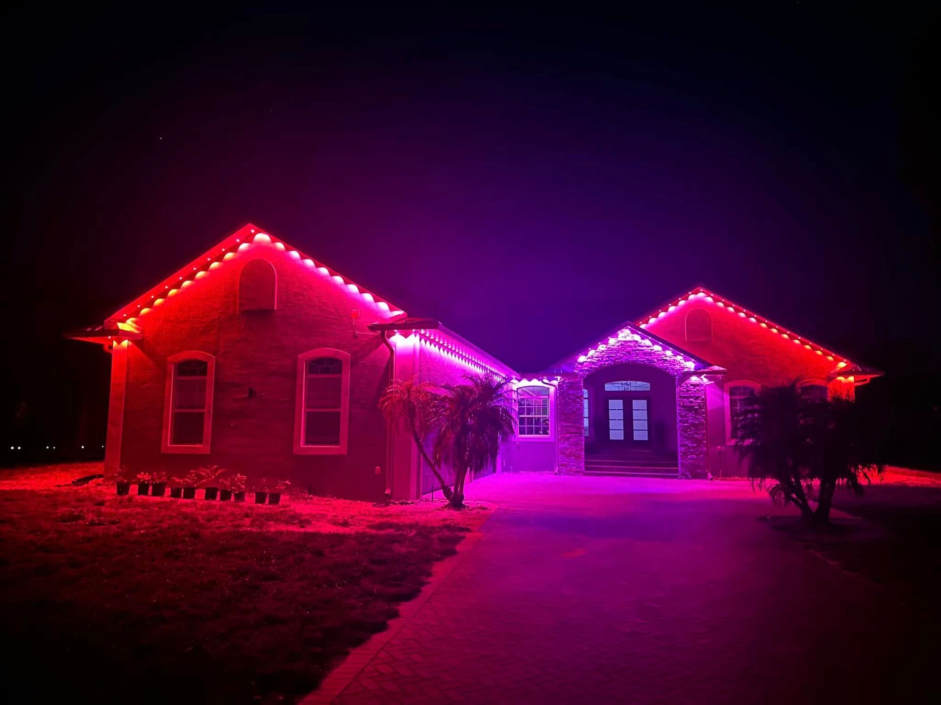 A house at night illuminated with vibrant red and purple string lights along the roofline and landscaping.