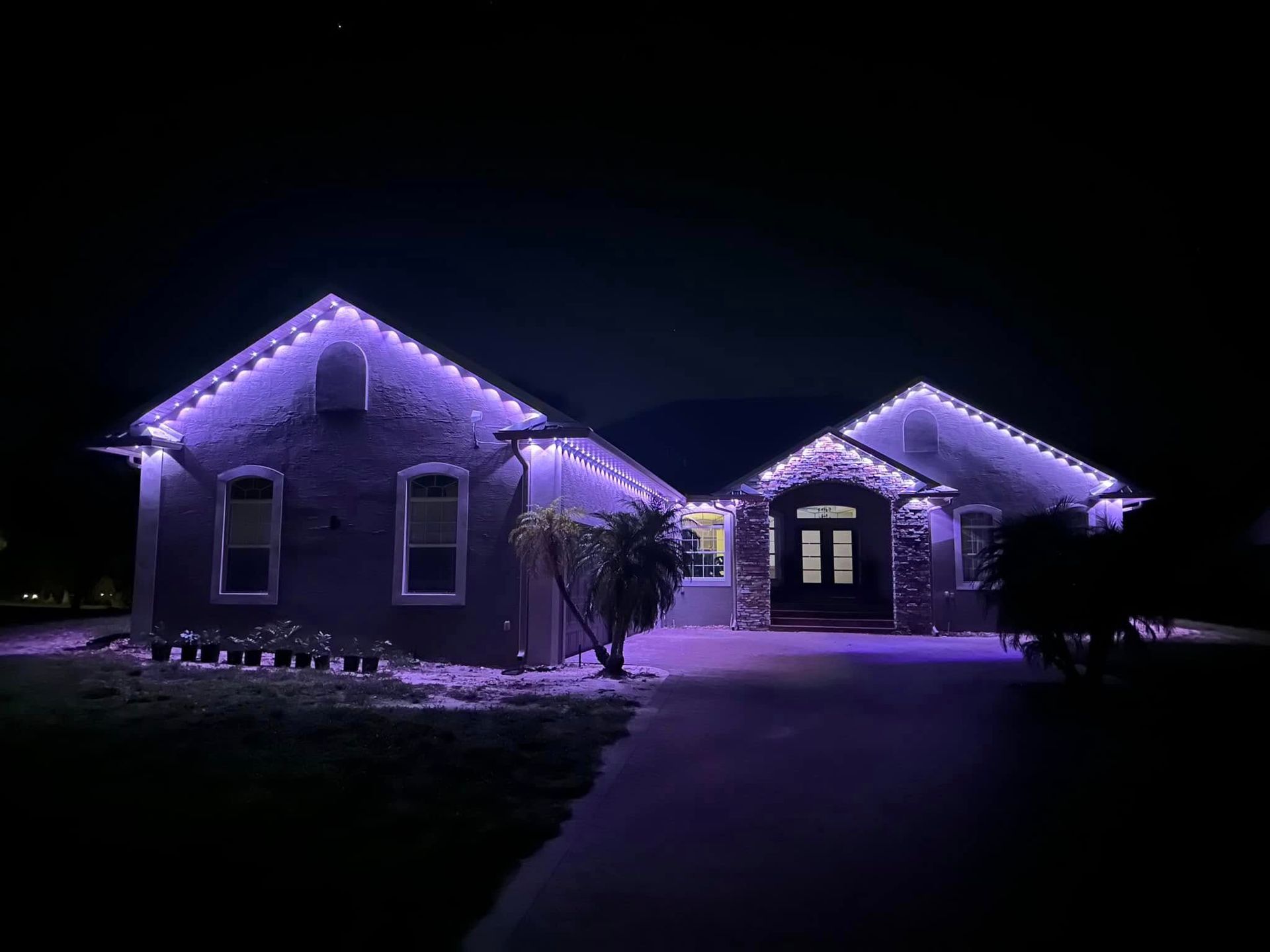 A house at night illuminated with purple roofline lighting, featuring a driveway and landscaping in the foreground.