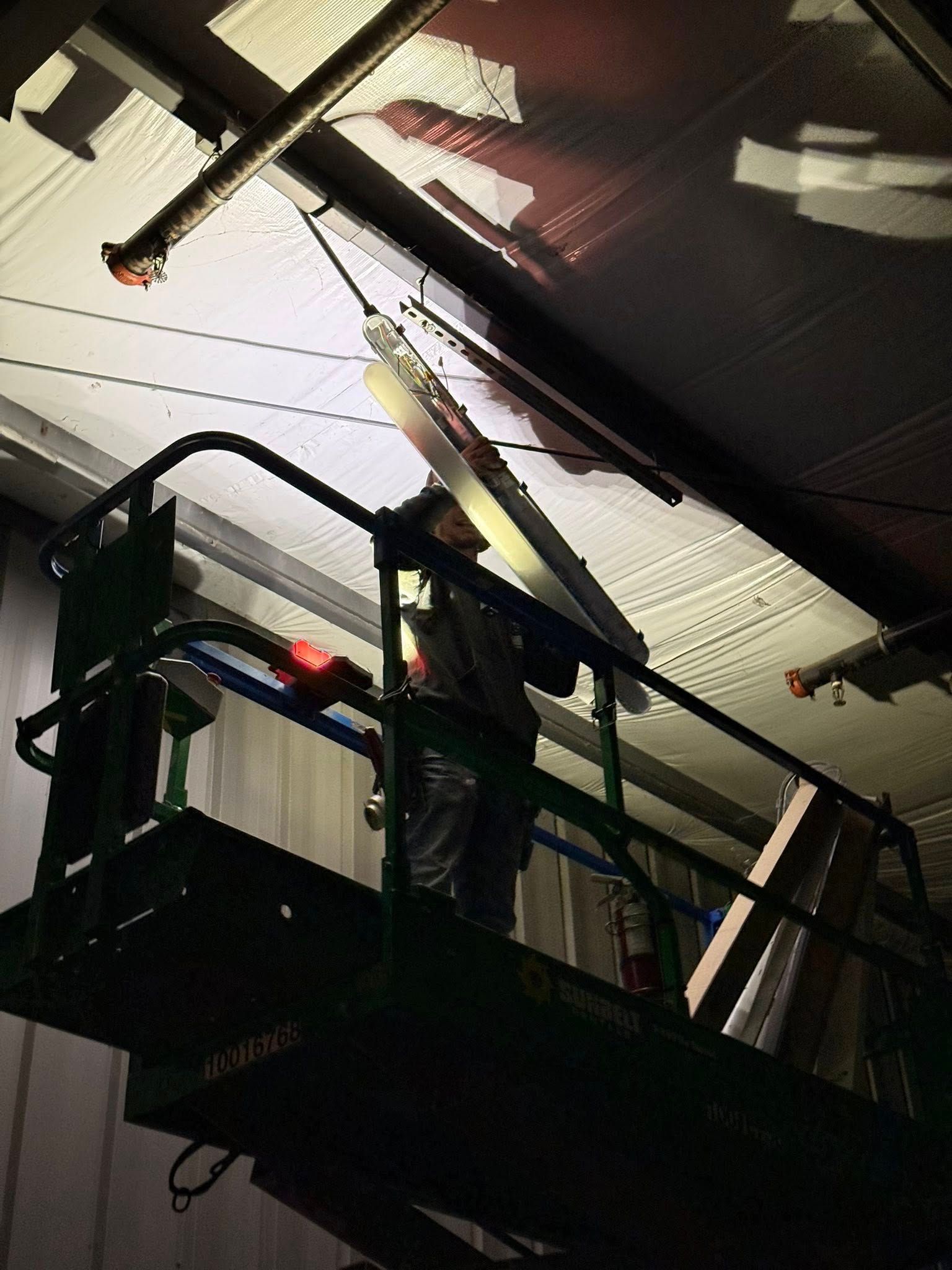 A worker on a scissor lift platform installs or services equipment mounted to a high ceiling in a dim, industrial space.