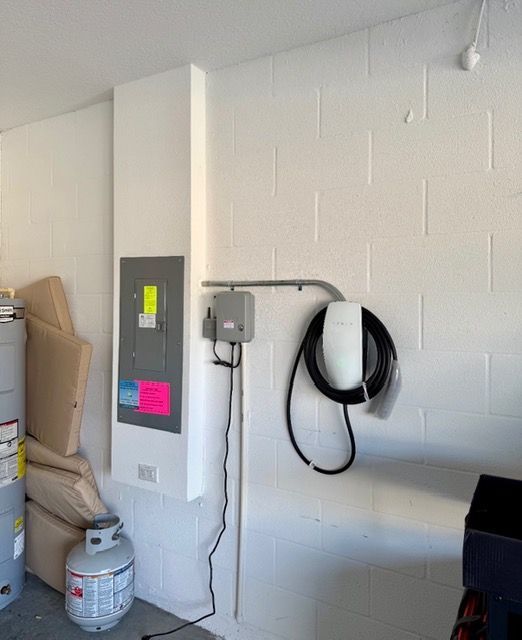 An electric vehicle wall charger and a gray disconnect box mounted to a white cinder block wall next to an electrical panel.