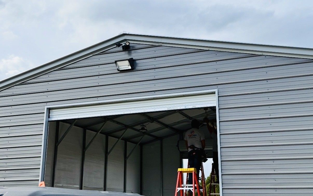 A worker stands on a red ladder inside an open grey metal building while performing repairs on the ceiling structure.