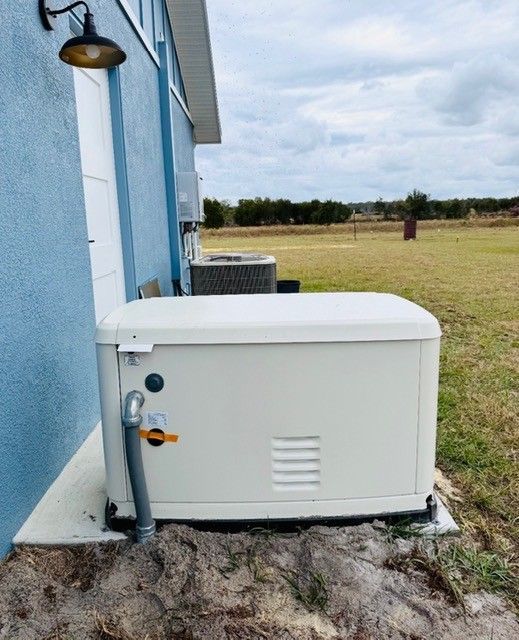 A white standby home generator sits on a concrete pad next to the blue exterior wall of a house with an open grassy field.