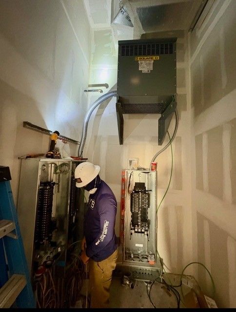 An electrician works on two open electrical breaker panels in a room with a wall-mounted heater above.