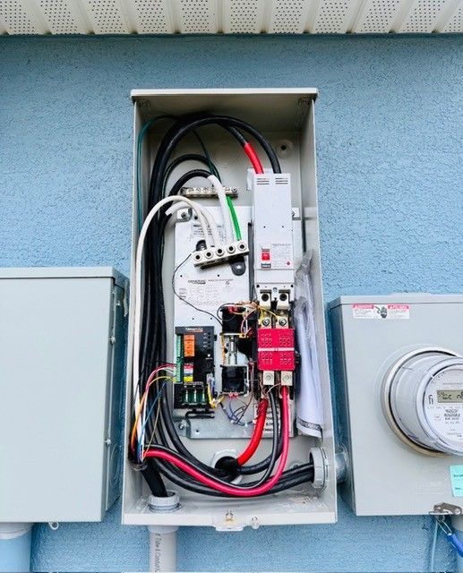 Open electrical service panel on a blue exterior wall with thick black and red wires, a circuit breaker, and a meter box.