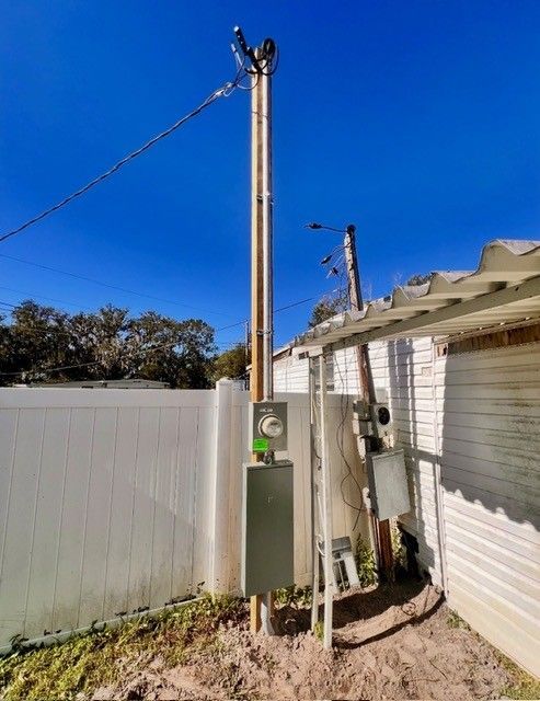 Two wooden utility poles with electrical meters and breaker boxes stand in a sandy yard next to a white fence and building.