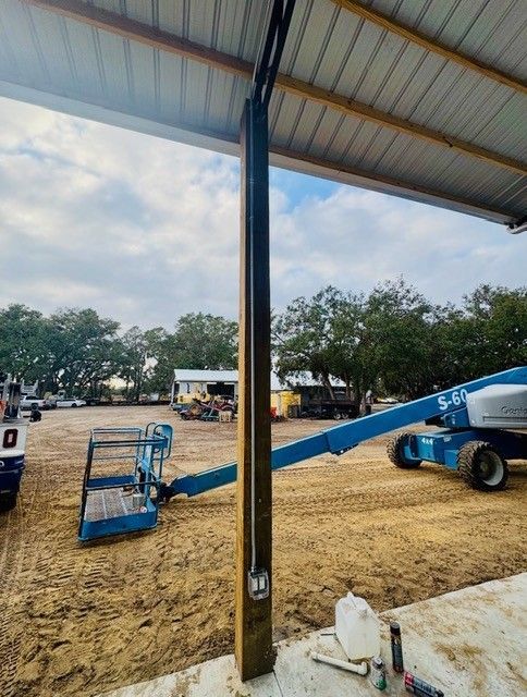 A blue industrial lift arm extends across a dirt lot, with a vertical black support beam in the foreground.