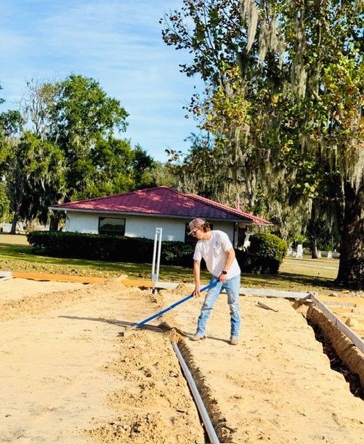 A person in a white shirt and jeans levels sandy ground with a tool in front of a house with a red metal roof.