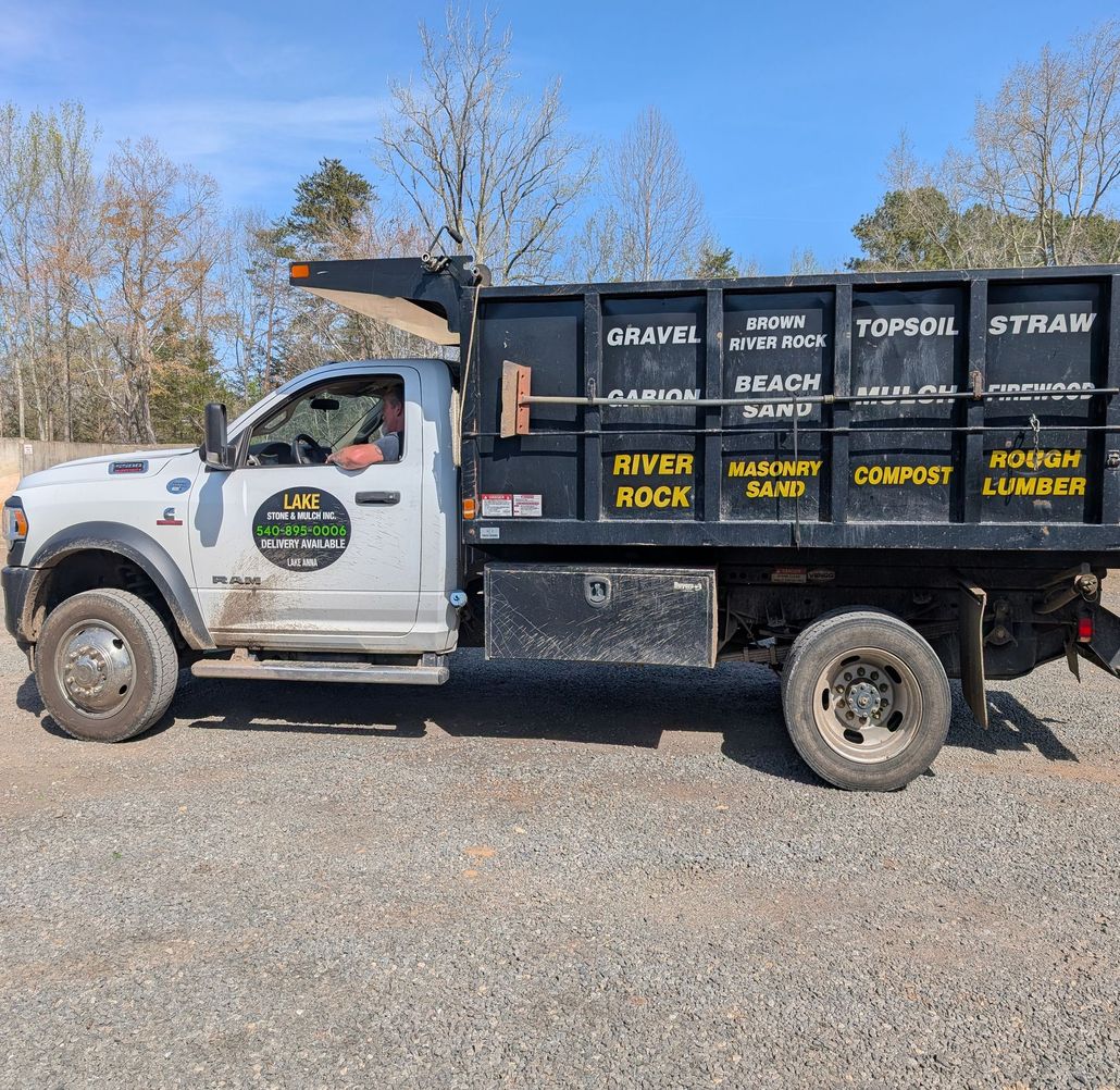 A white dump truck parked on a gravel lot features a black bed labeled with various landscaping materials for sale.