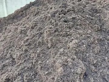 A large, textured pile of dark brown organic mulch or compost sitting against a light-colored concrete wall.