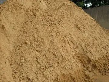 A large pile of light brown, coarse construction sand outdoors with a blurred wall and tree in the background.