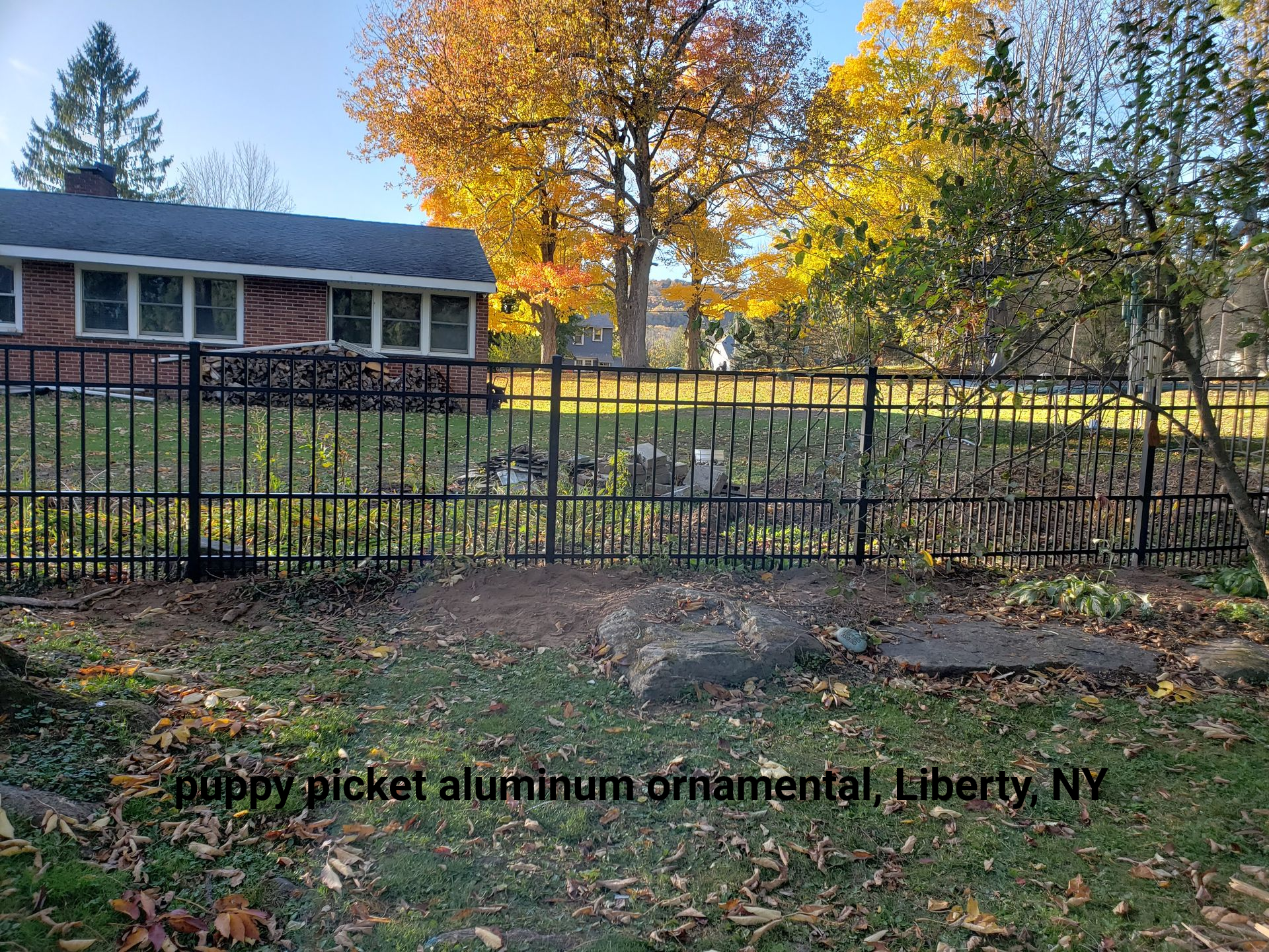 Black picket aluminum ornamental fence in front of a yard with fall foliage and a brick house in Liberty, NY.