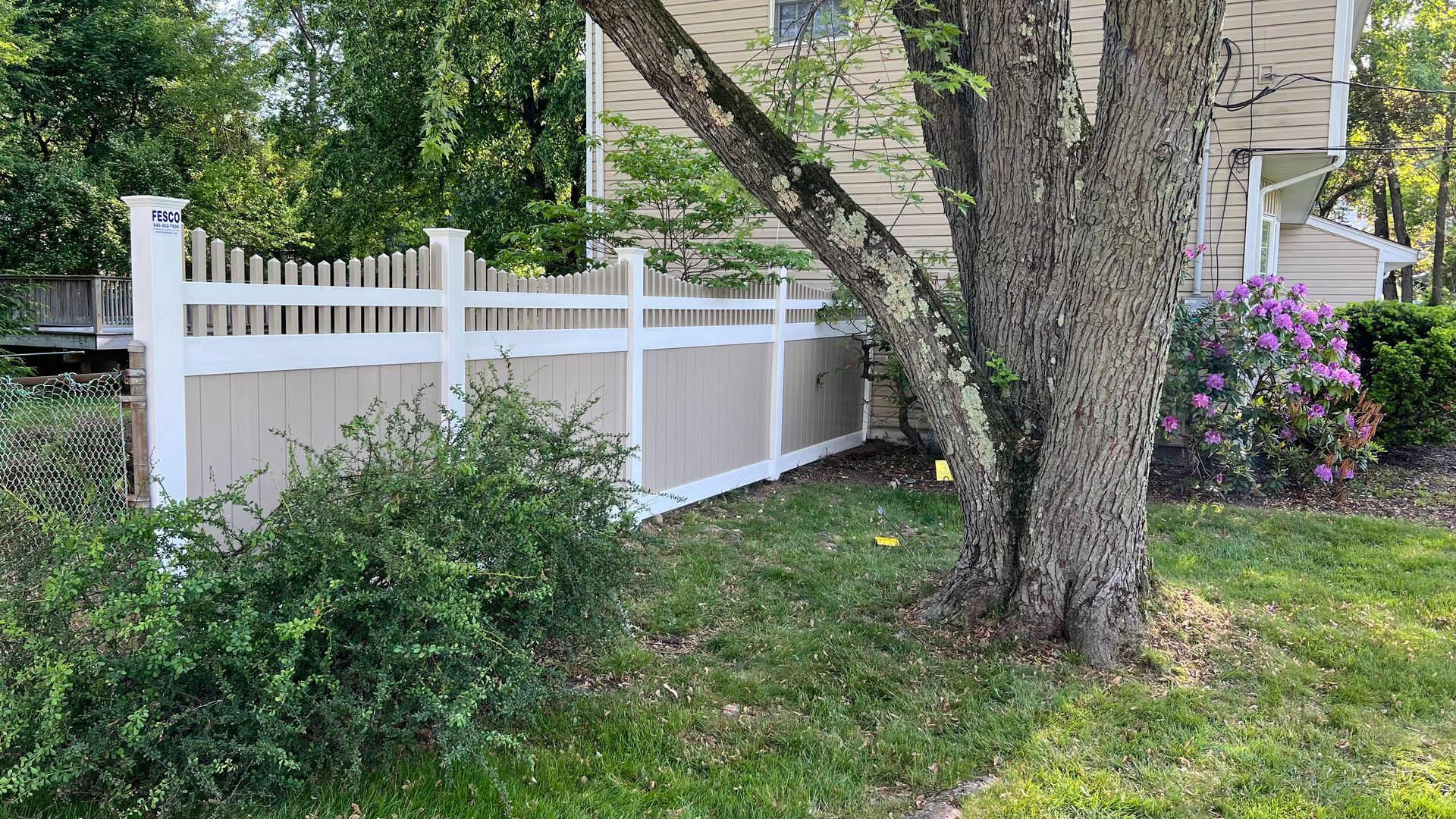 White picket fence with tan panels, next to a tree, lawn, and house.