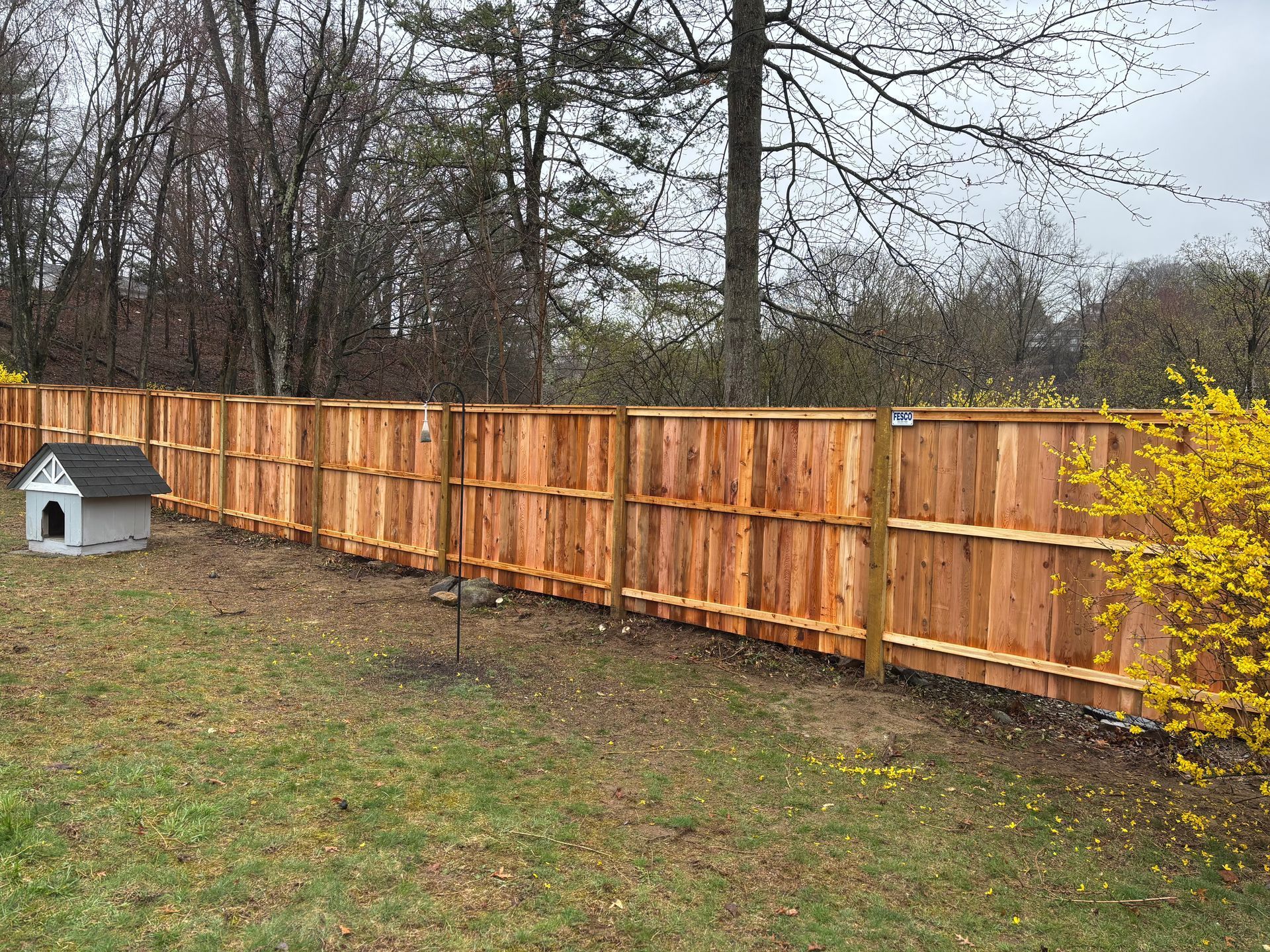 Wooden fence in a backyard with a dog house, surrounded by trees and grass, on an overcast day.