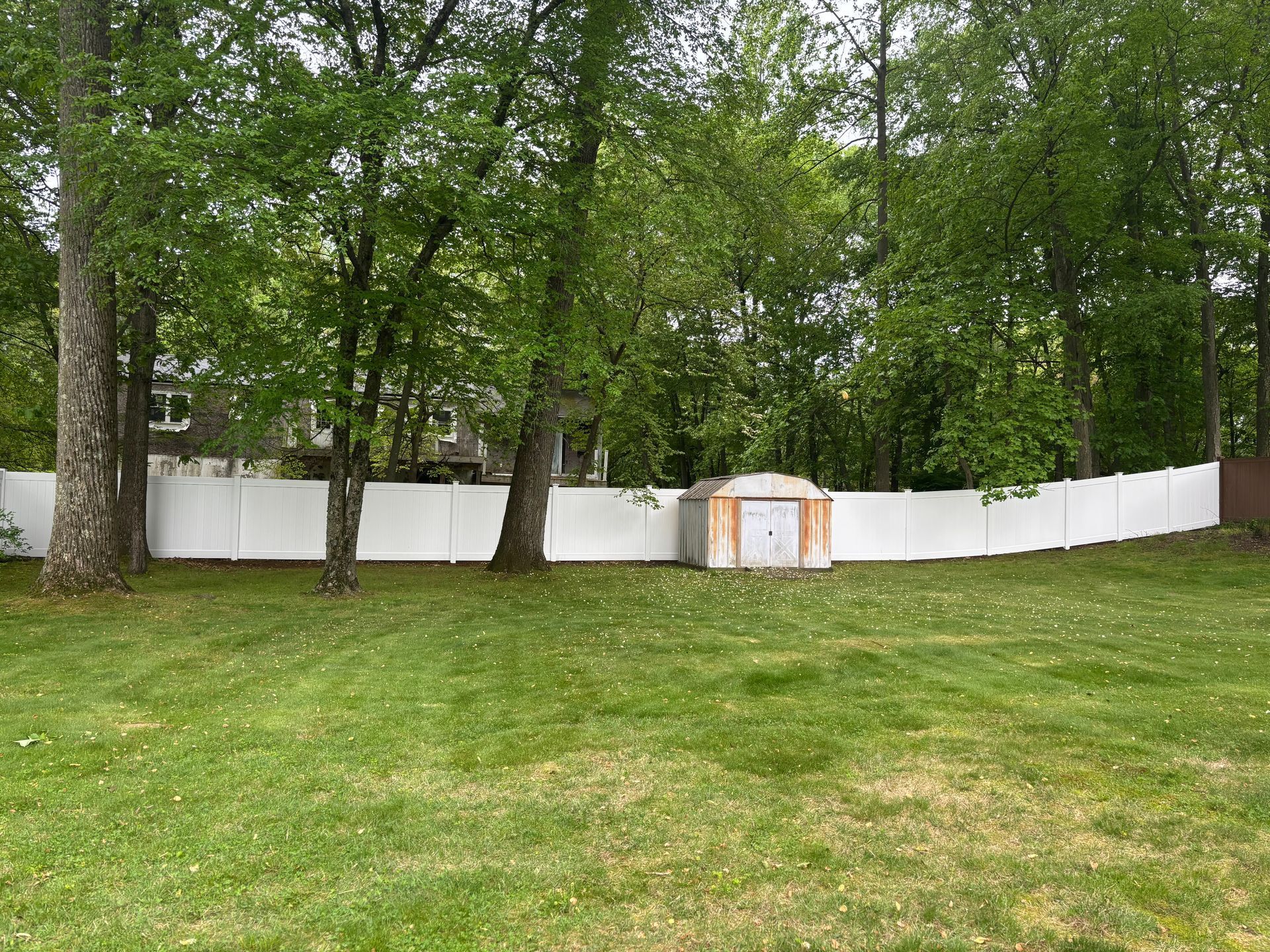White fence encloses a backyard with green grass and trees; a small shed sits inside the fence.