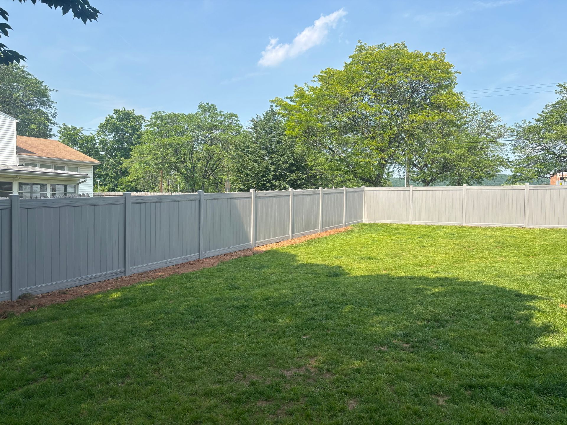 Gray vinyl fence encloses a green lawn under a blue sky, trees in the background.