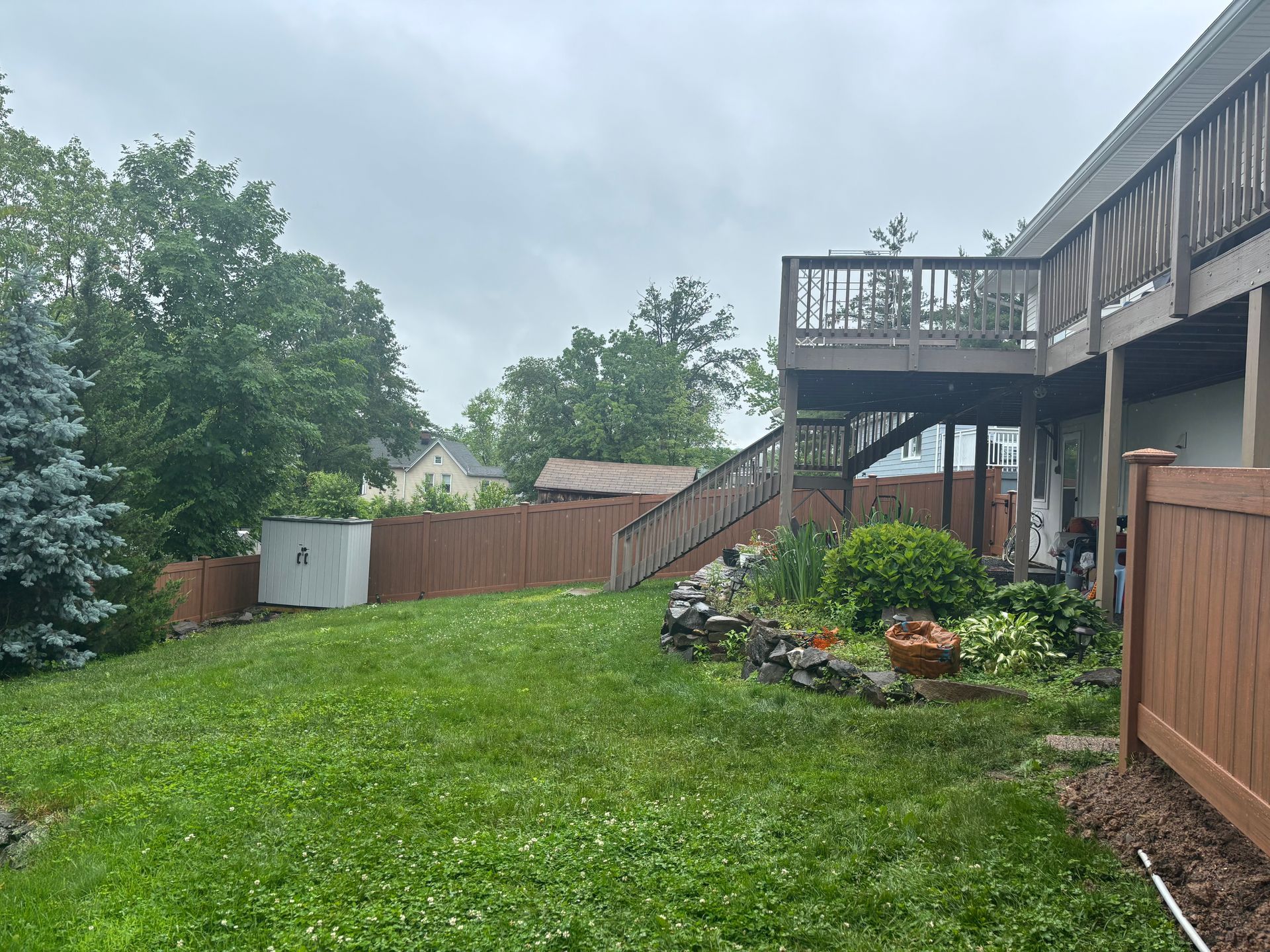 Backyard with brown deck, fence, shed, and green grass on a cloudy day.