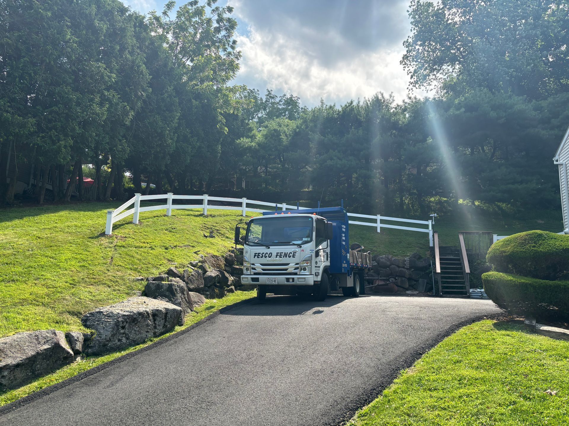 Truck parked on a driveway in front of a white fence and stairs, next to green grass and trees.