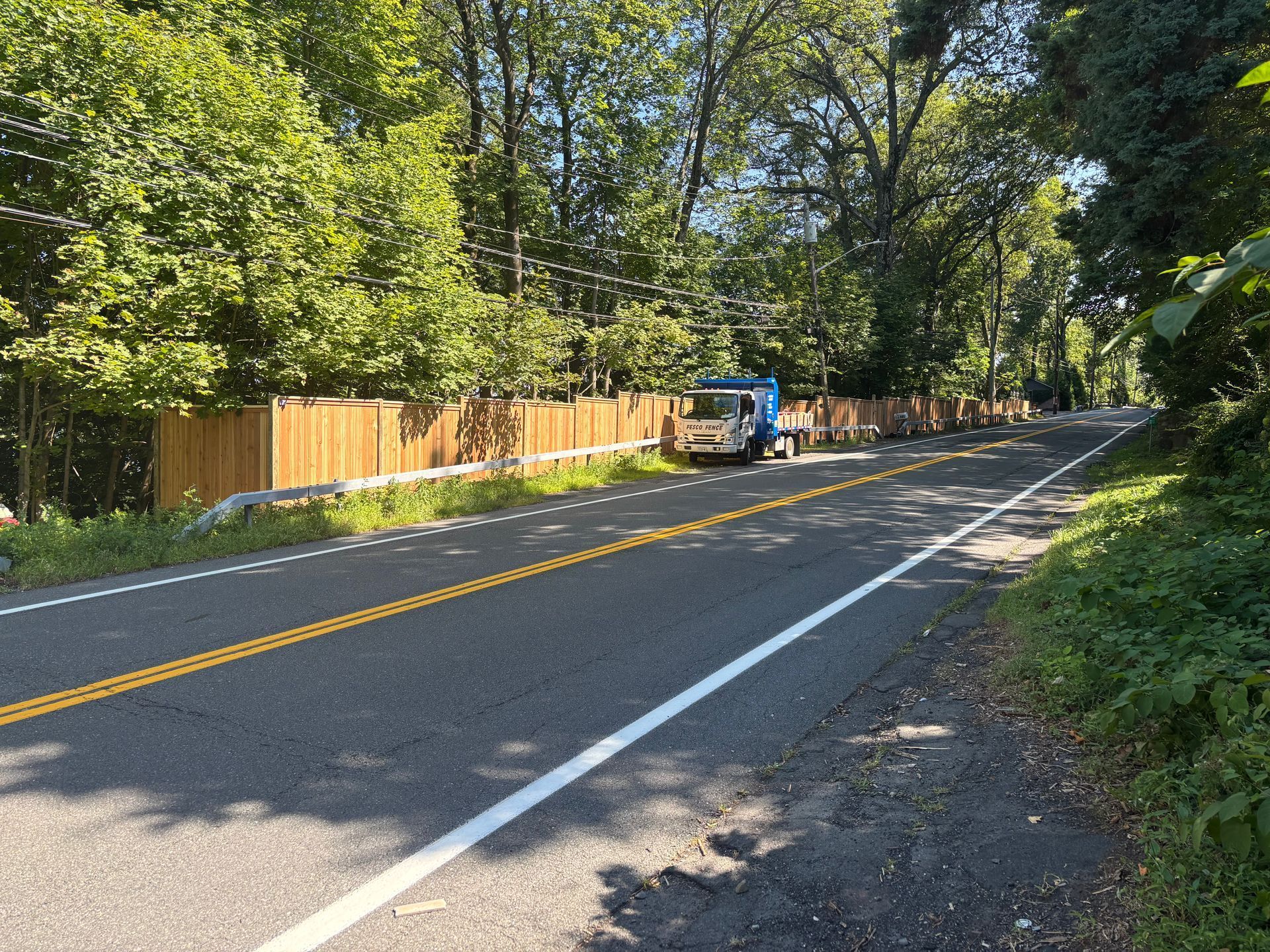 Truck parked on a road lined with a wooden fence and trees, on a sunny day.