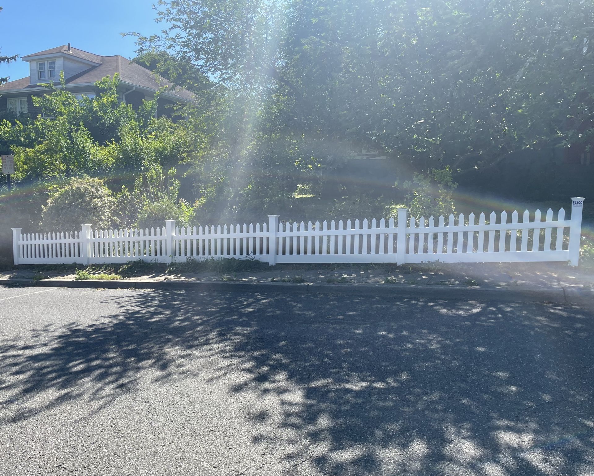 White picket fence along a paved road, sunlight streams through trees, a house in background.