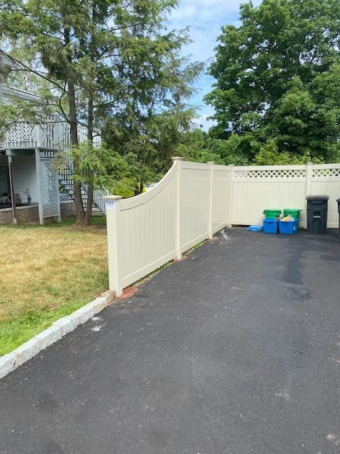 A driveway bordered by a tan fence, a grassy area, and trees, with blue and green bins.