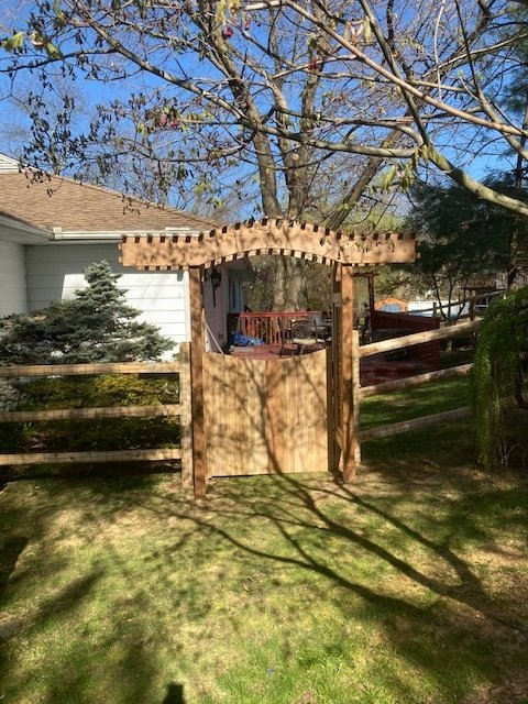 Wooden arbor with gate in a yard; sunlight creates shadows on the grass and structure.