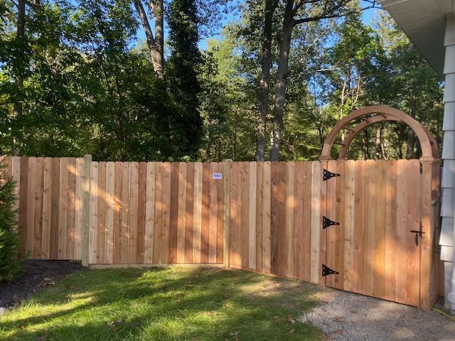 Wooden fence with gate, arched top, set in a yard with trees.