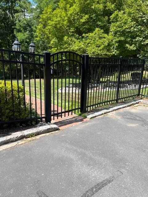 Black metal gate in a yard with trees, brick pathway, and asphalt driveway.