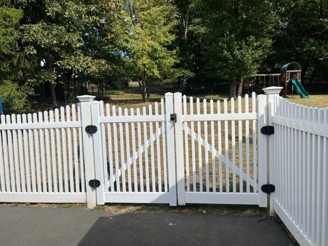 White picket fence with double gate, in a sunny yard with trees and a playground in the background.